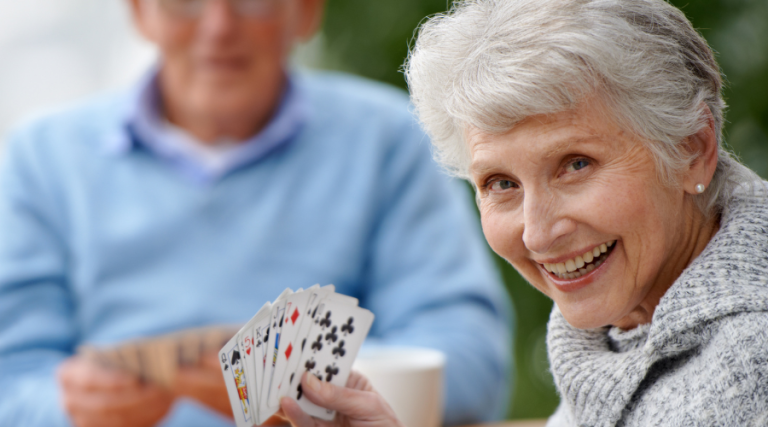 Two seniors playing bridge, in the foreground a woman looks at the camera and smiles, holding up her bridge hand. To illustrate and article titled Cognitive benefits of bridge and moderate strategic gaming