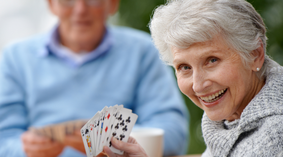 Two seniors playing bridge, in the foreground a woman looks at the camera and smiles, holding up her bridge hand. To illustrate and article titled Cognitive benefits of bridge and moderate strategic gaming
